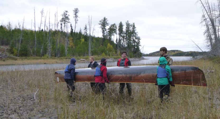 Six students carry a North Canoe, a large freighter canoe, near a body of water framed by trees. 